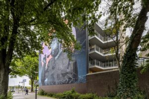 Muurschildering van een man met een pijp op een flatgebouw aan de Prins Hendrikkade, deels omgeven door bomen.